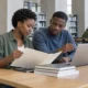 Deux jeunes adultes consultent un dossier et un ordinateur dans une bibliothèque.